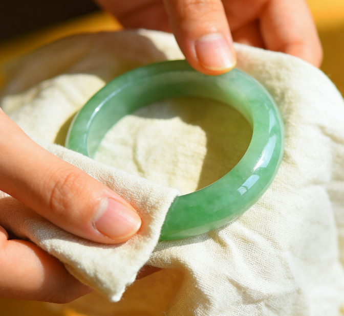 Hands gently cleaning a green nephrite jade bracelet with a soft microfiber cloth in natural daylight